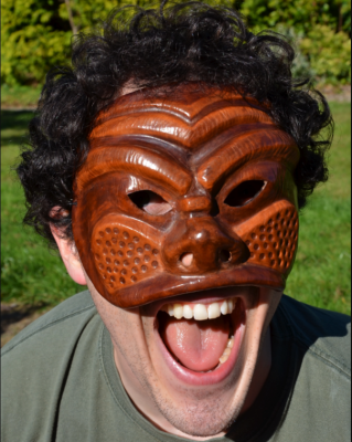 this is an image of a man wearing a commedia mask - it is brown, intricately carved wood that covers the face down to the nose and cheeks. It has holes for the eyes and little breathing holes for the nose.